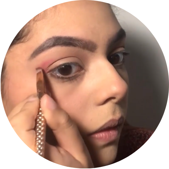 Rosto de uma mulher se maquiando. Ela segura com a mão direita um pincel onde passa uma sombra rosa no seu olho.