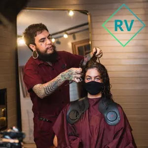 Homem usando barba longa, com pente e tesoura cortando o cabelo ondulado de uma mulher que está sentada na cadeira do salão.