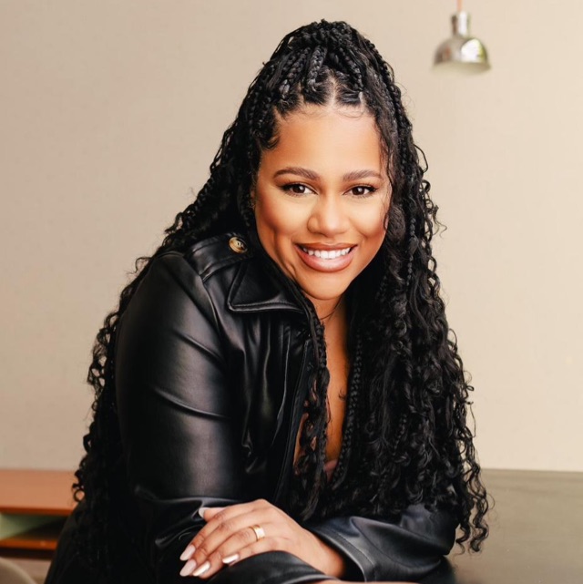 Mulher negra de cabelos longos, com uma parte presa bem ao alto. Ela veste uma jaqueta preta e está sorrindo para frente.