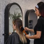 Mulher em pé, usando um jaleco preto realiza escovação no cabelo da cliente que está sentada em um salão profissional.
